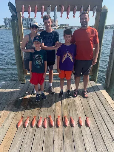 Four people pose on a dock beside rows of freshly caught fish, with water and boats in the background.