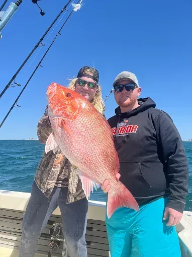 Two people on a boat holding a large pink fish against a bright blue sky and ocean