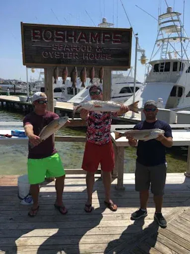 Three people posing with a fish on a dock in front of an oyster house sign.