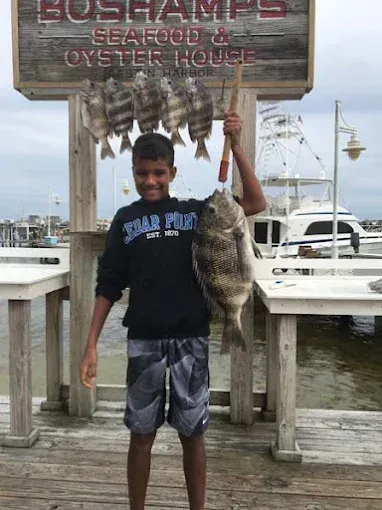 Man holding a large fish on a dock beside a seafood house sign and boats