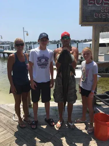 Four people standing on a wooden pier by the water, two holding drinks, one in a red cap.
