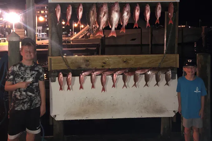 Two boys posing beside a large display of freshly caught fish at night.