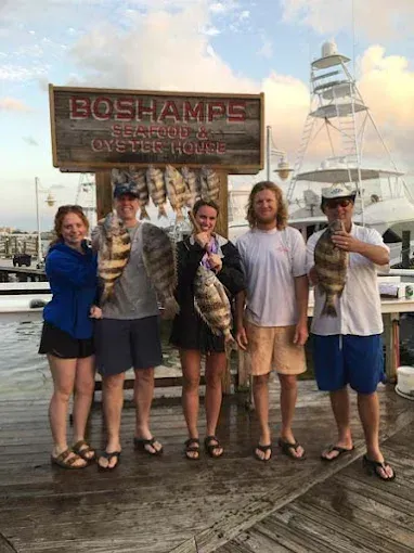 Four people holding large fish beside a BOSHAMPS Oyster House sign on a dock, with a boat in the background