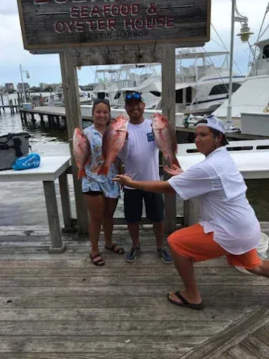 Three people on a dock posing with two large fish at a seafood restaurant marina.