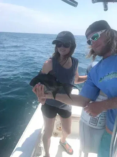 Two people on a boat holding a fish over the ocean, with blue water and sky in the background.