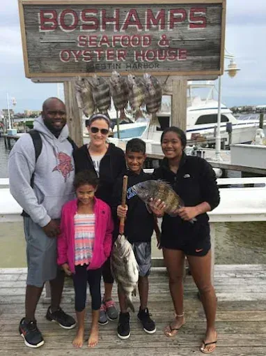 Group posing at Boshamps Seafood & Oyster House dock, one holding a fish, with boats in the background