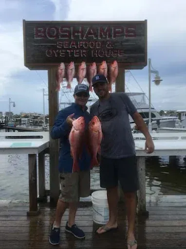 Two men holding large fish in front of a dockside seafood house sign.