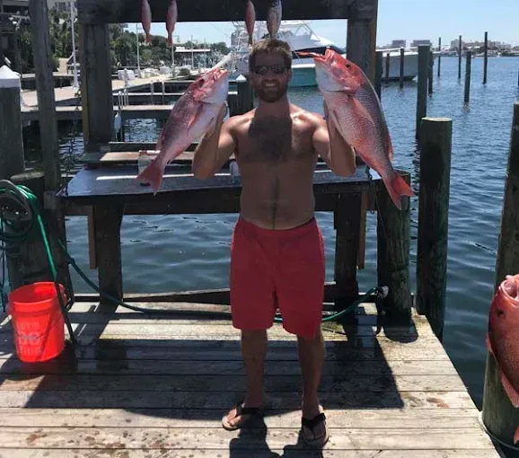 Man on a dock holding two large fish, with water and boats in the background.