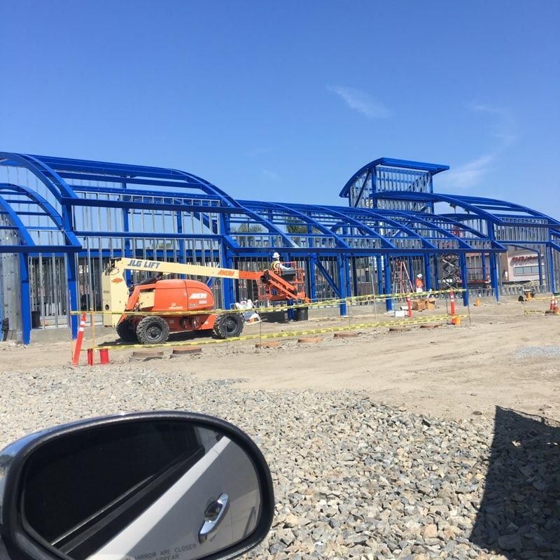 Blue steel frame building under construction with orange lift and cones on a dirt site under a clear sky