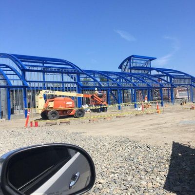 Blue steel building under construction with orange lift trucks and traffic cones on a dirt site