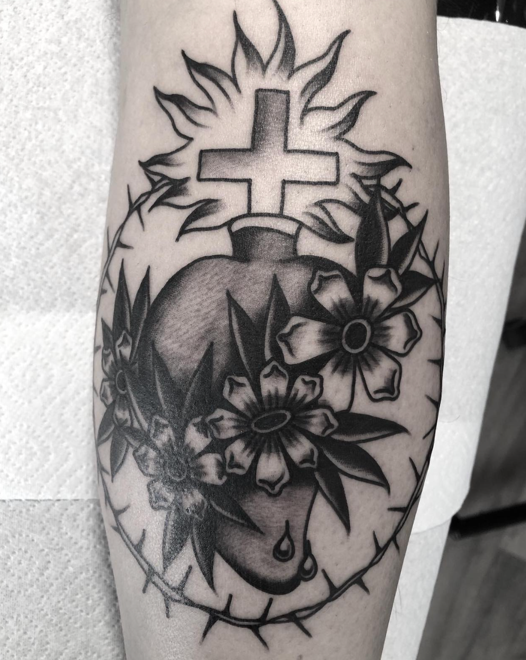 Black-and-gray tattoo of a heart with flowers and a cross above it on a person’s forearm