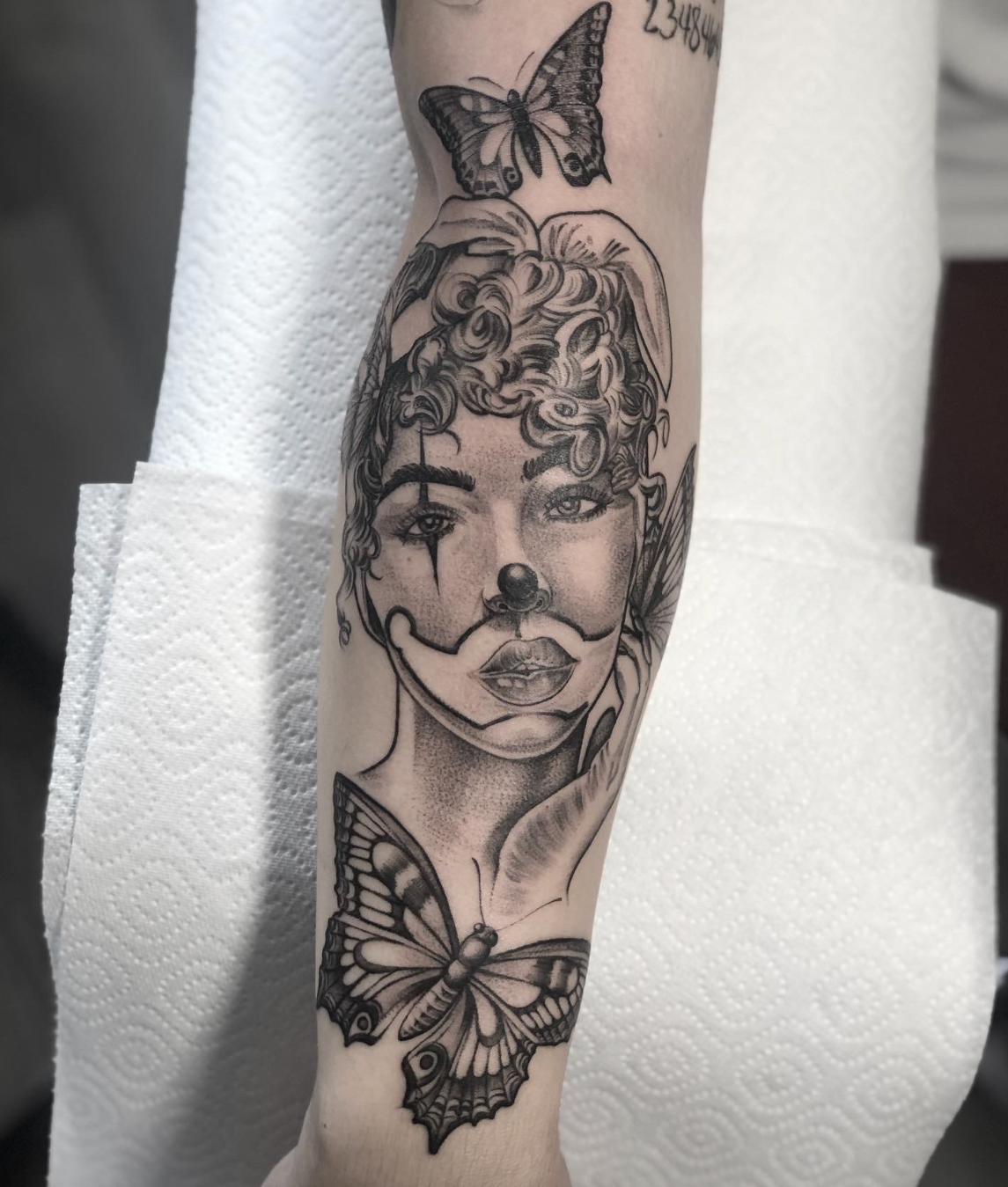 Black-and-gray tattoo of a face with butterfly details on a forearm, with flowers and a butterfly near the wrist