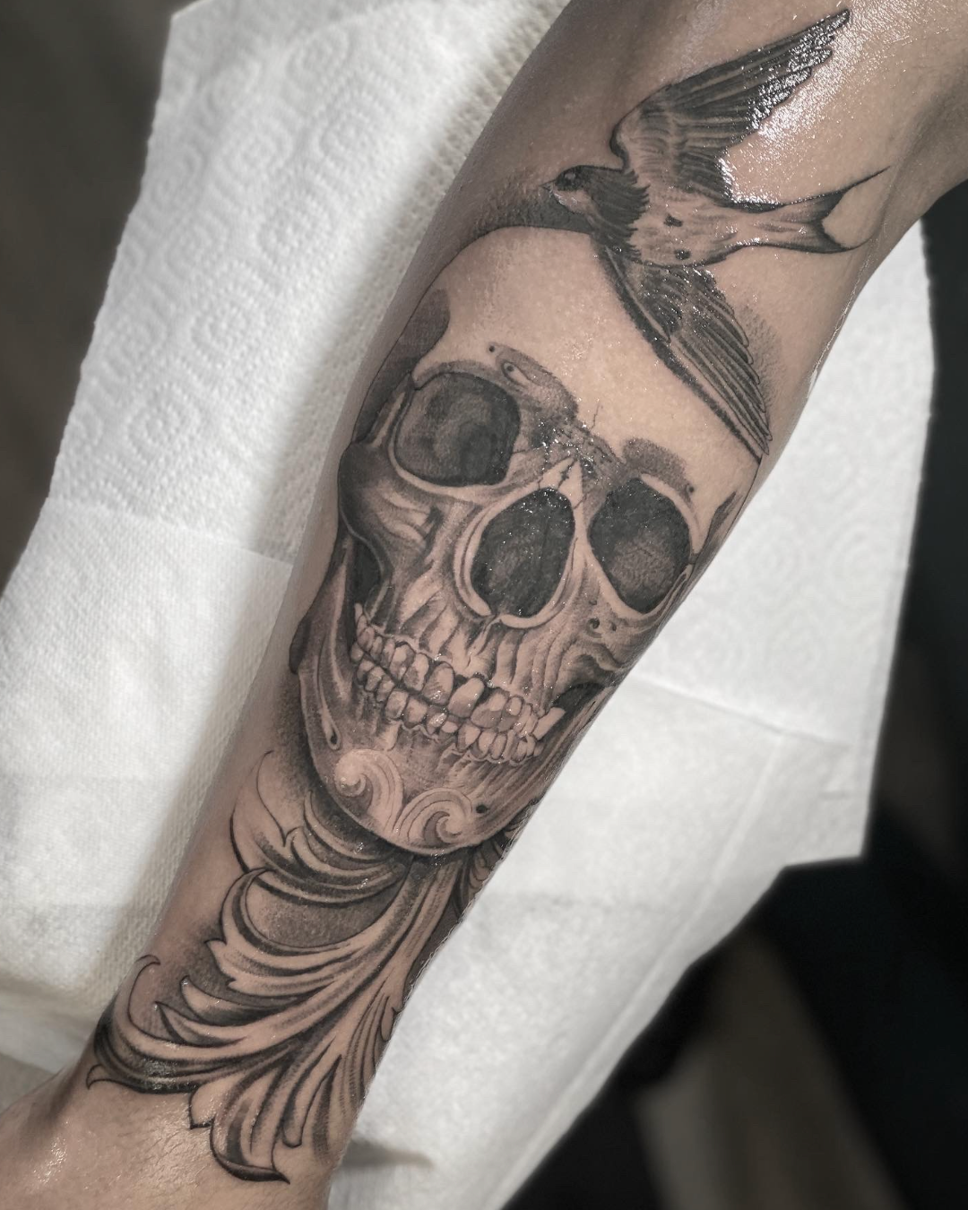 Black-and-gray skull tattoo with bird and floral scrollwork on a forearm.