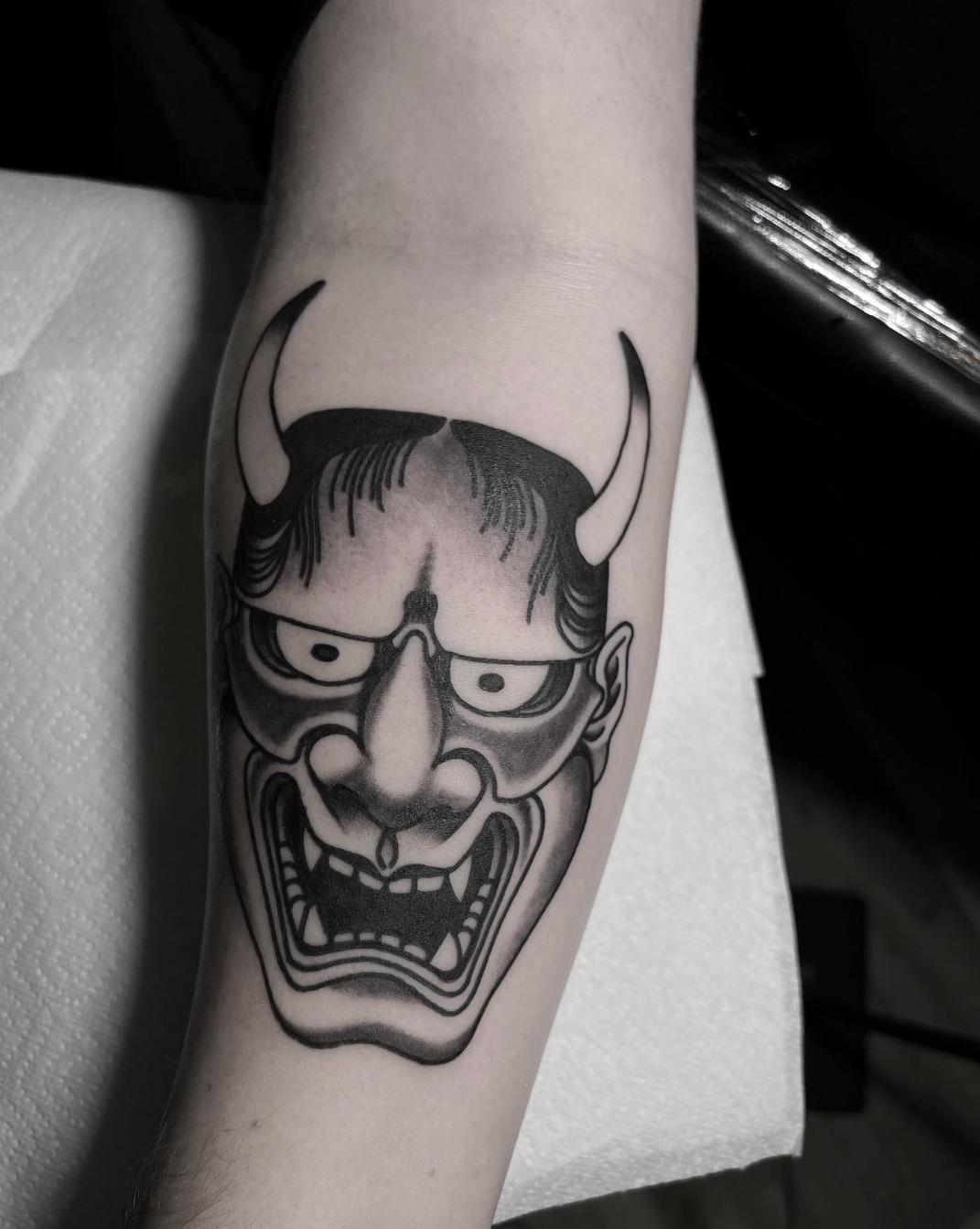 Black-and-gray horned oni mask tattoo on a forearm, with an angry expression and sharp teeth