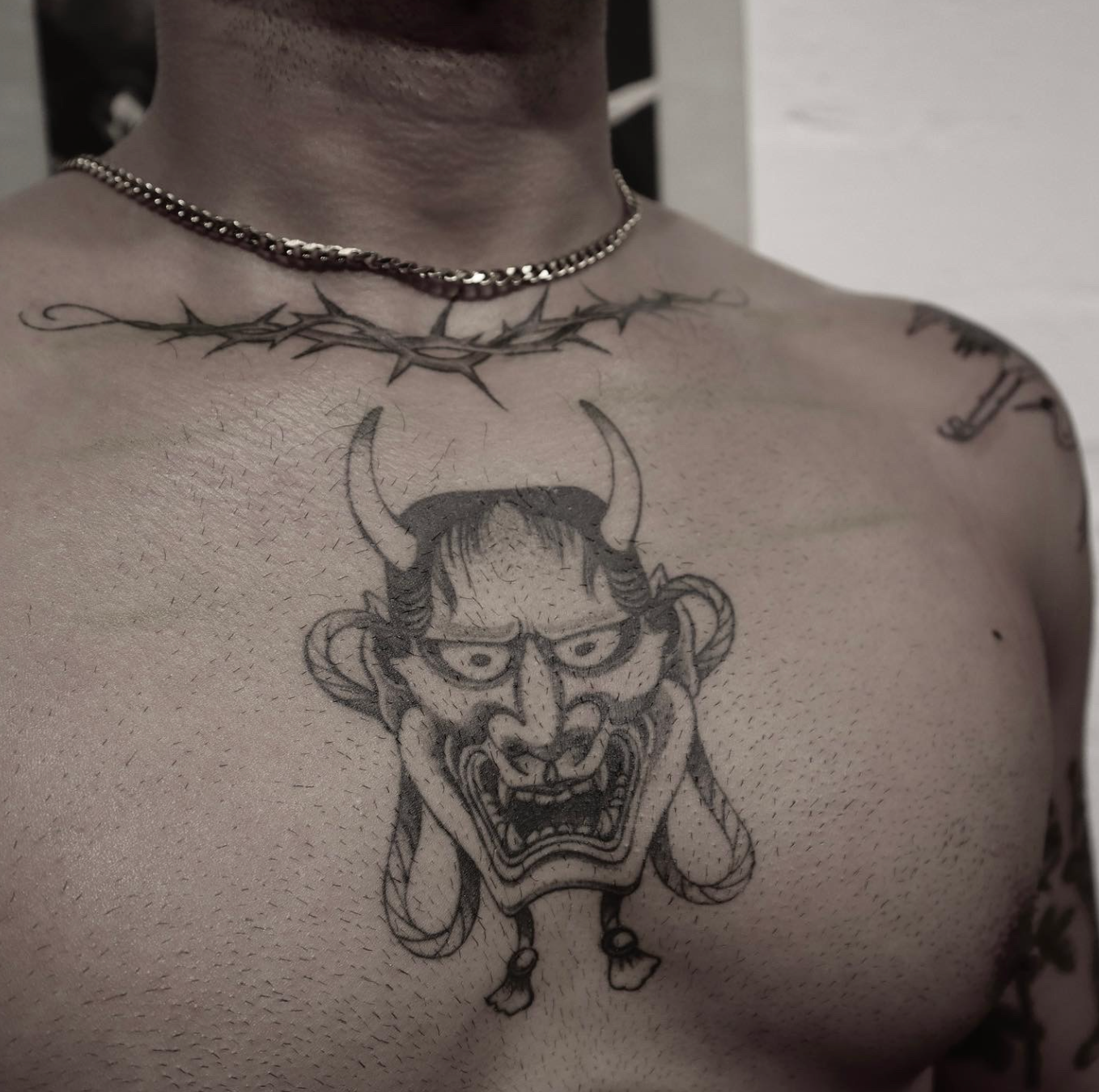 Bare-chested person wearing a necklace, with a dark horned face tattoo centered on the chest