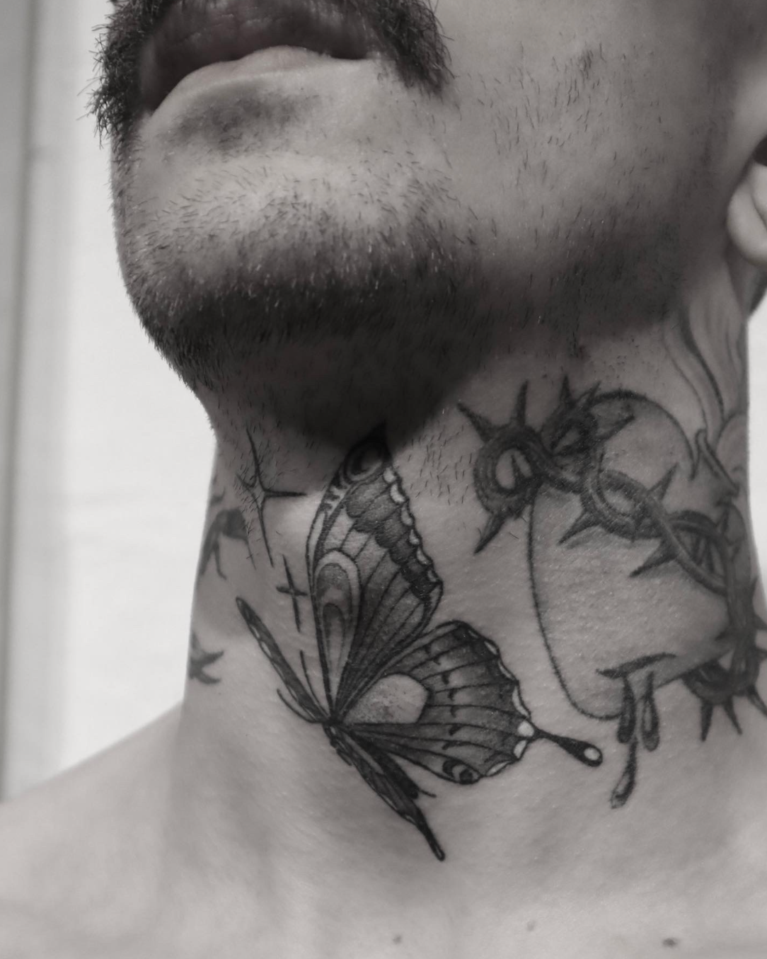 Black-and-white close-up of a man's neck with butterfly and beetle tattoos beneath his jawline