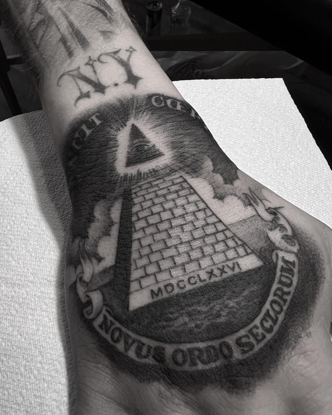 Forearm tattoo of an all-seeing pyramid with a triangle, stars, and a banner reading “NOVUS ORDO SECLORUM.”