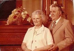 Two people pose in front of a floral arrangement in a wood-paneled room.
