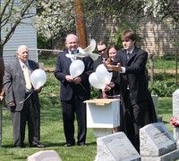 Four people in formal attire release white balloons at a graveside memorial ceremony.