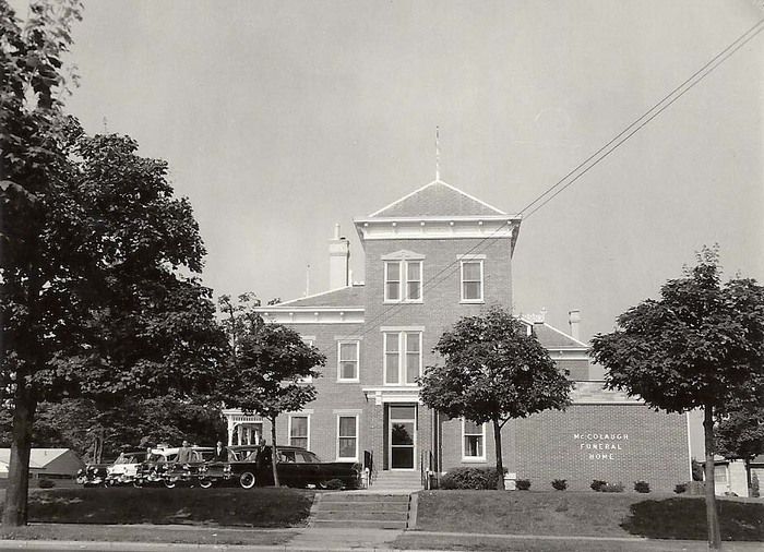 Historic brick building with a small tower, trees in front, and a sign on the lawn