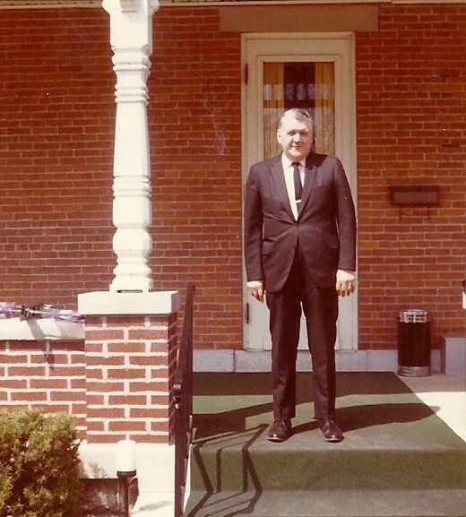 Man in a dark suit standing on a porch outside a brick building, in front of a white door.
