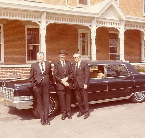 Three men in suits posing beside a black vintage car in front of a brick building.
