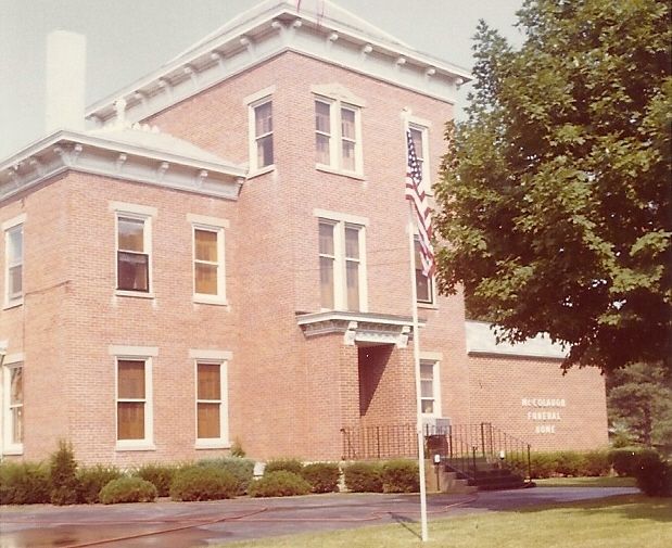 Brick building with white trim and shrubs in front, viewed from a lawn with a tree on the right.