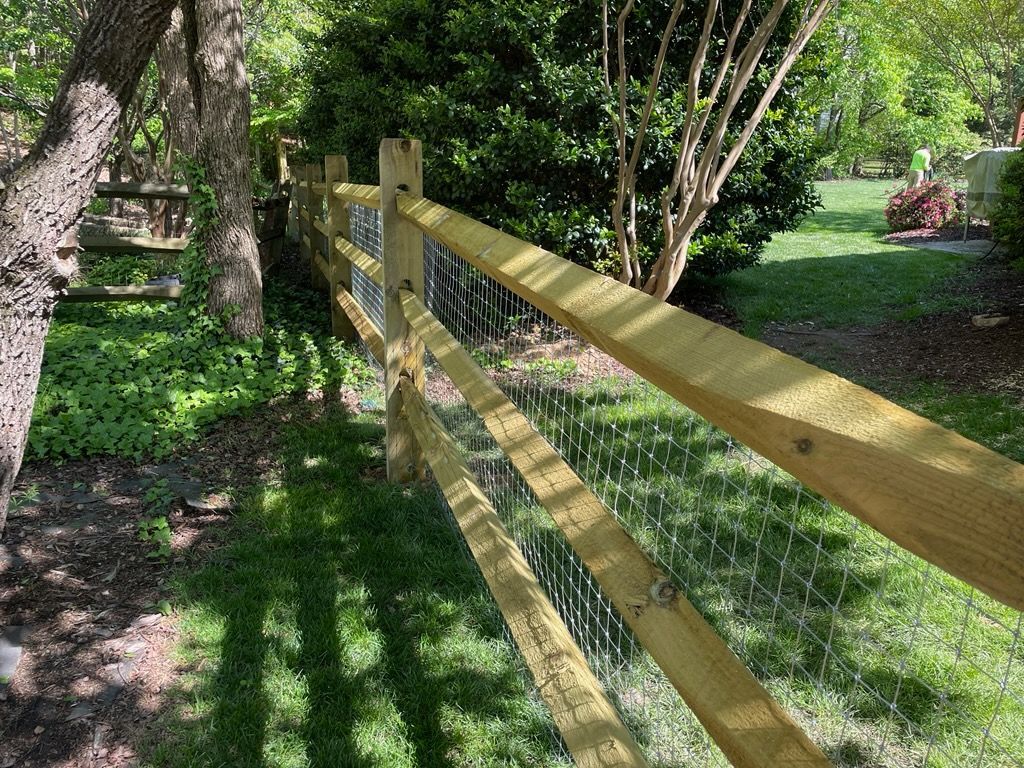Wooden split-rail fence with wire mesh in a grassy yard, trees in the background.