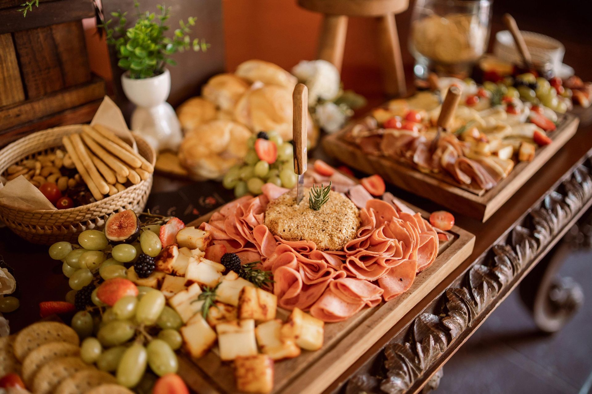 A banquet table features wooden boards arranged with various cheeses, cured meats, grapes, bread, and crackers.