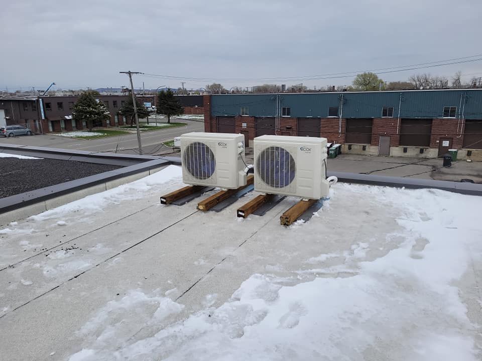 Deux unités de climatisation blanches sur des supports en bois, sur un toit plat parsemé de plaques de neige.