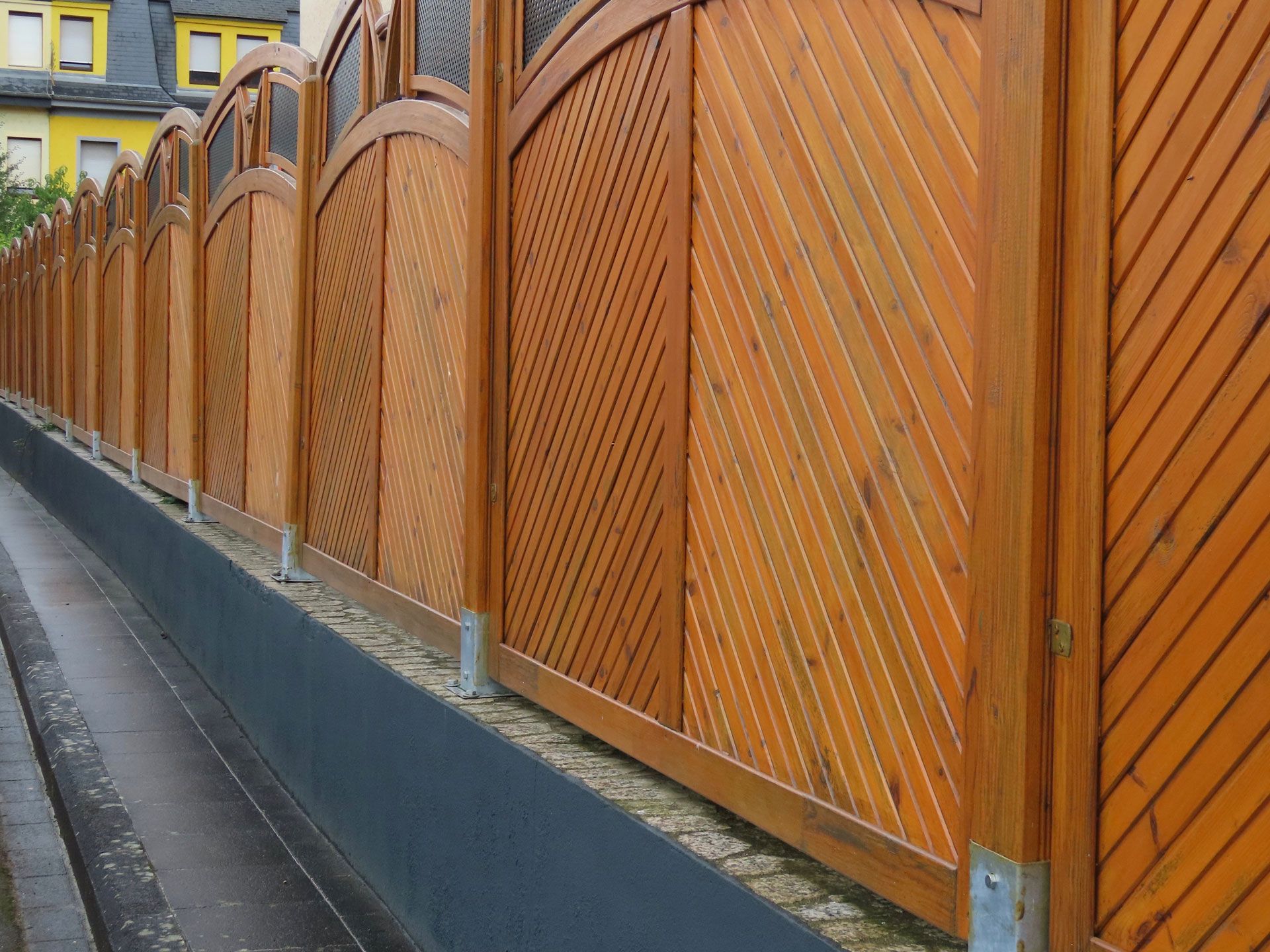 Orange wooden fence along a sidewalk beside a wet street and yellow building.