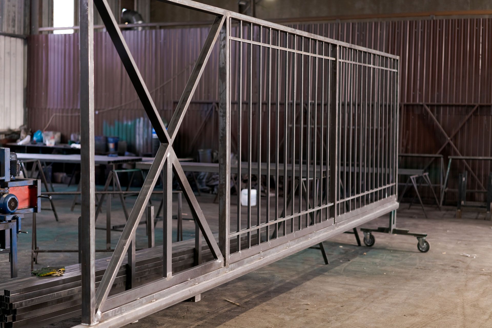 Large metal gate or railing frame in a workshop, with black cross-bracing and wheels on the floor.