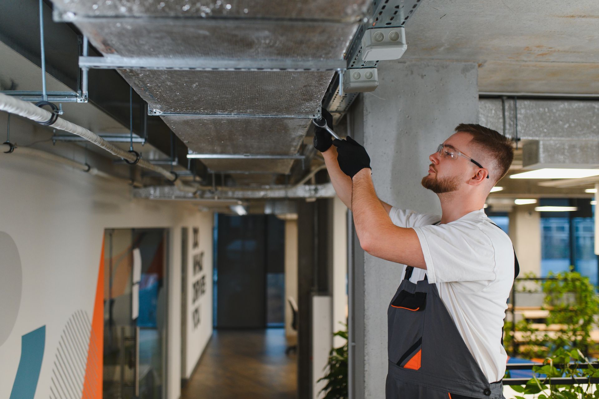 Worker in white shirt and black overalls inspecting overhead ductwork in a modern office corridor.