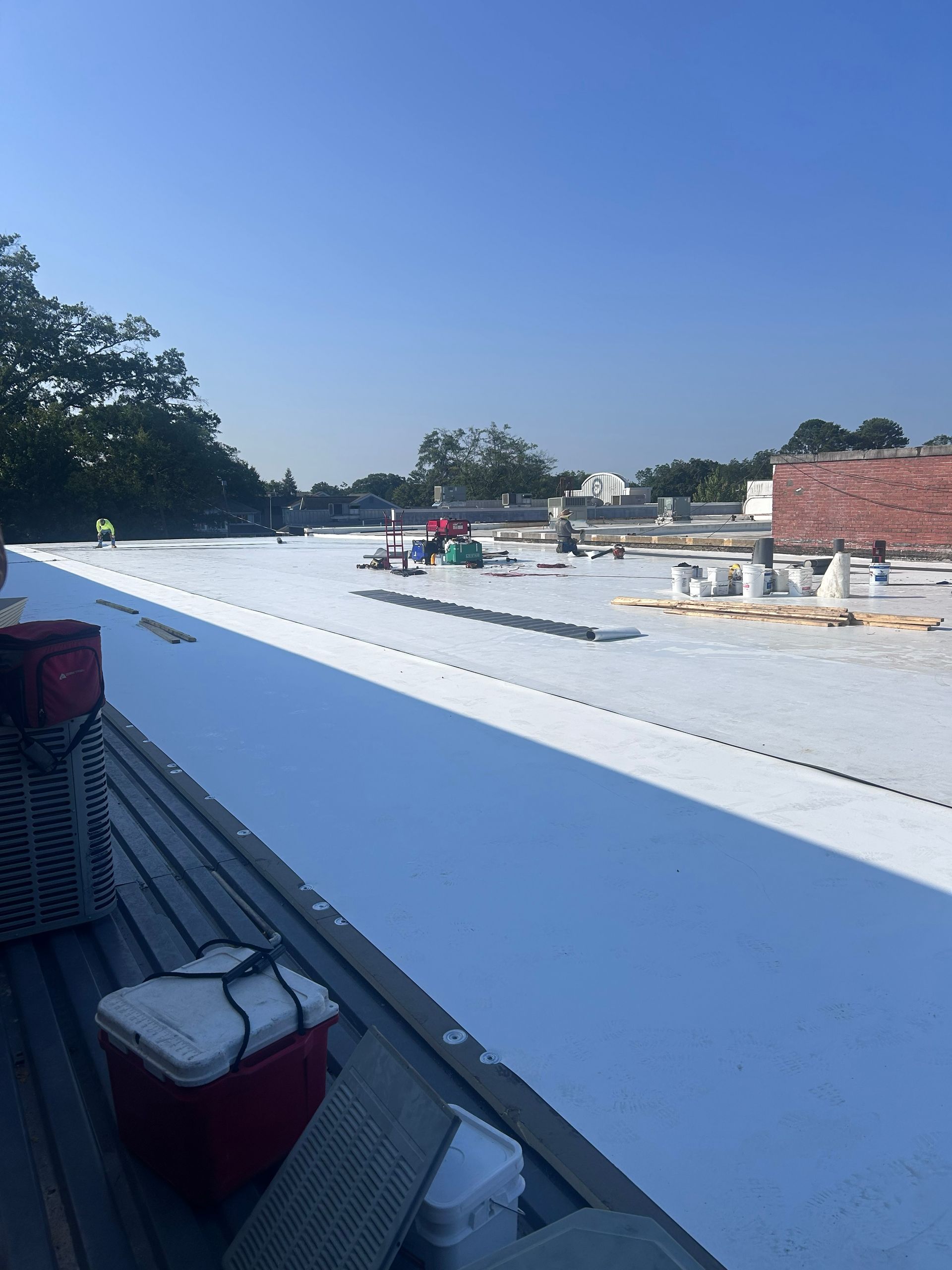 White roof under construction with workers, materials, and trees under a clear blue sky