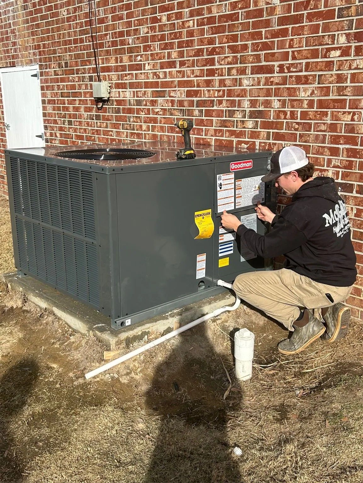 Technician kneels beside outdoor HVAC unit, inspecting controls against a brick wall.