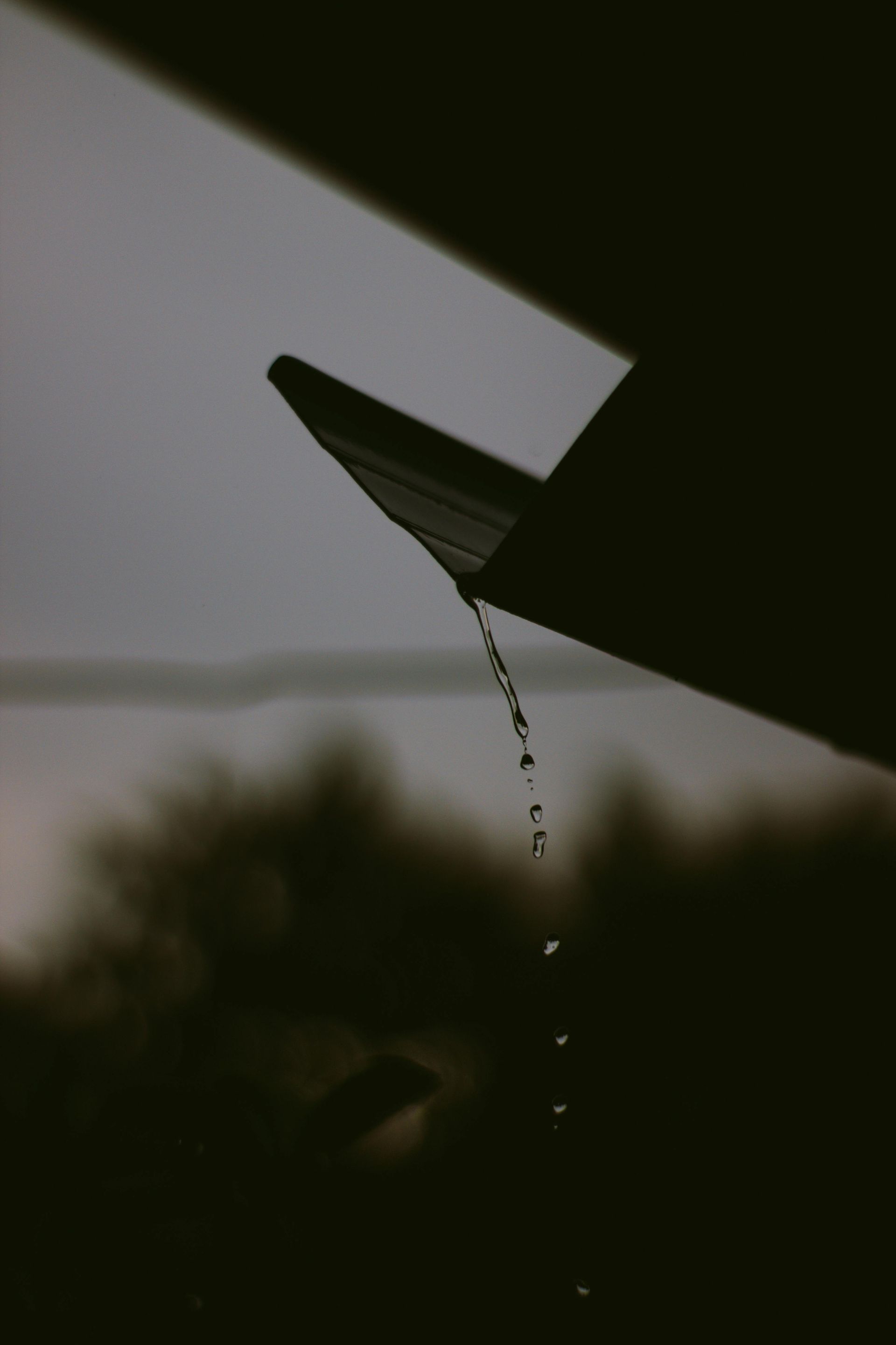 Rainwater drips from the gutter of a dark, angular roofline against a blurred, overcast background.