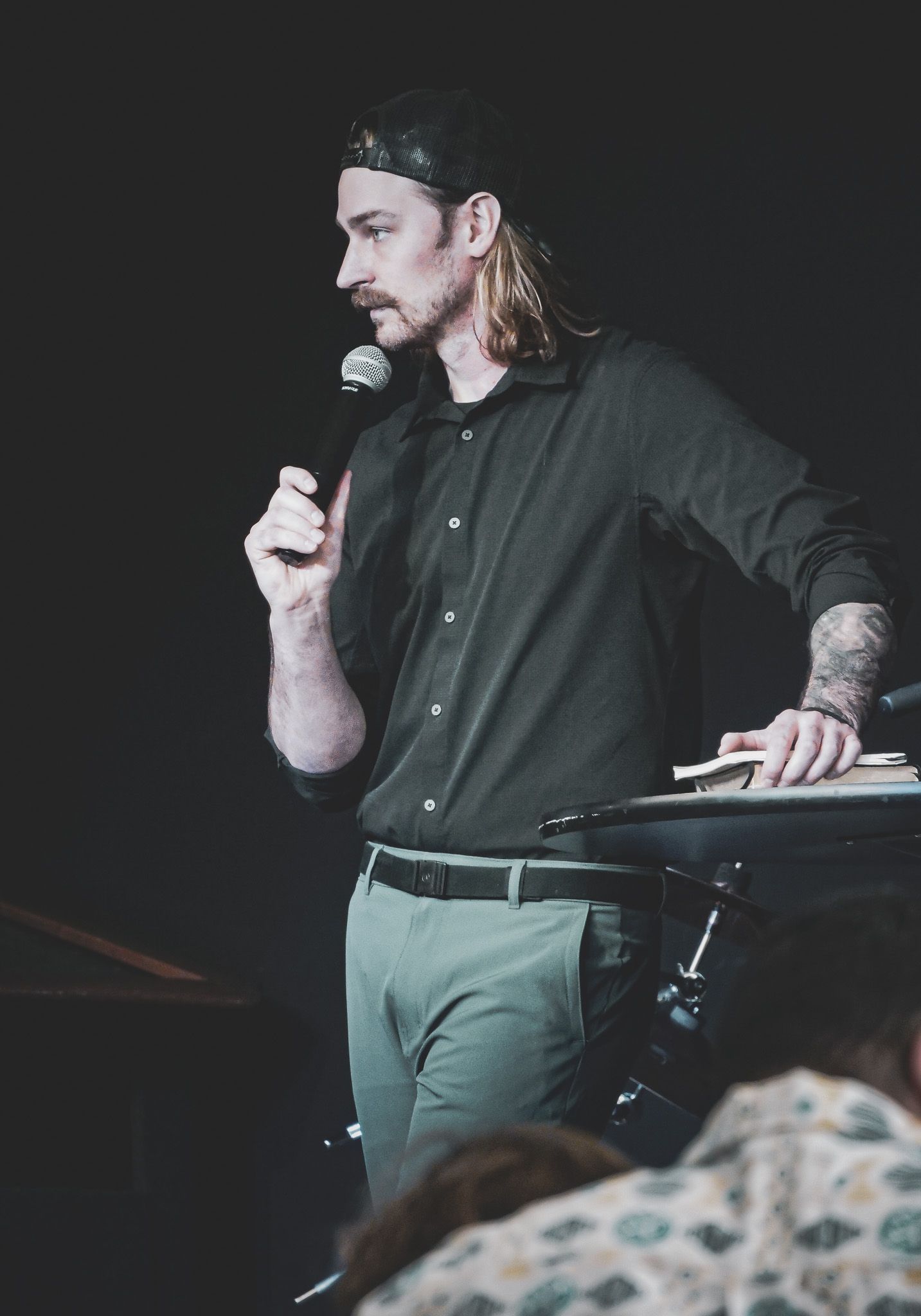 Man with mustache and long hair, holding microphone, speaking on stage, wearing green shirt and pants.