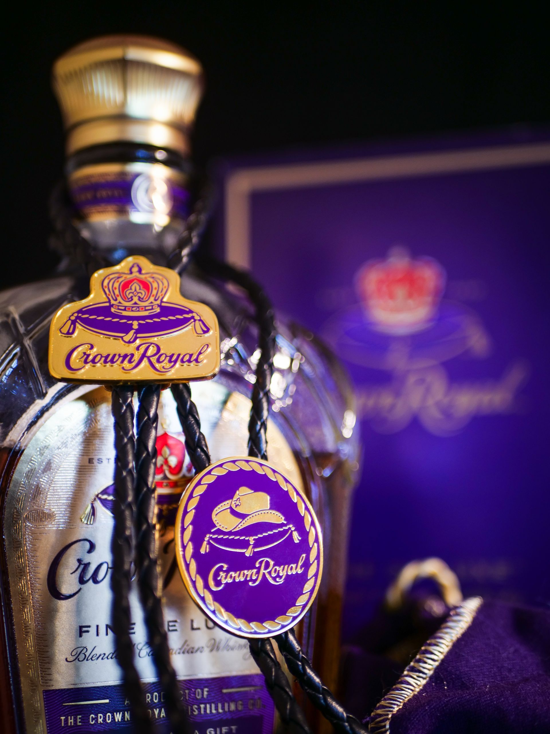 Crown Royal bottle with purple and gold label and a leather bolo tie, against a purple background.