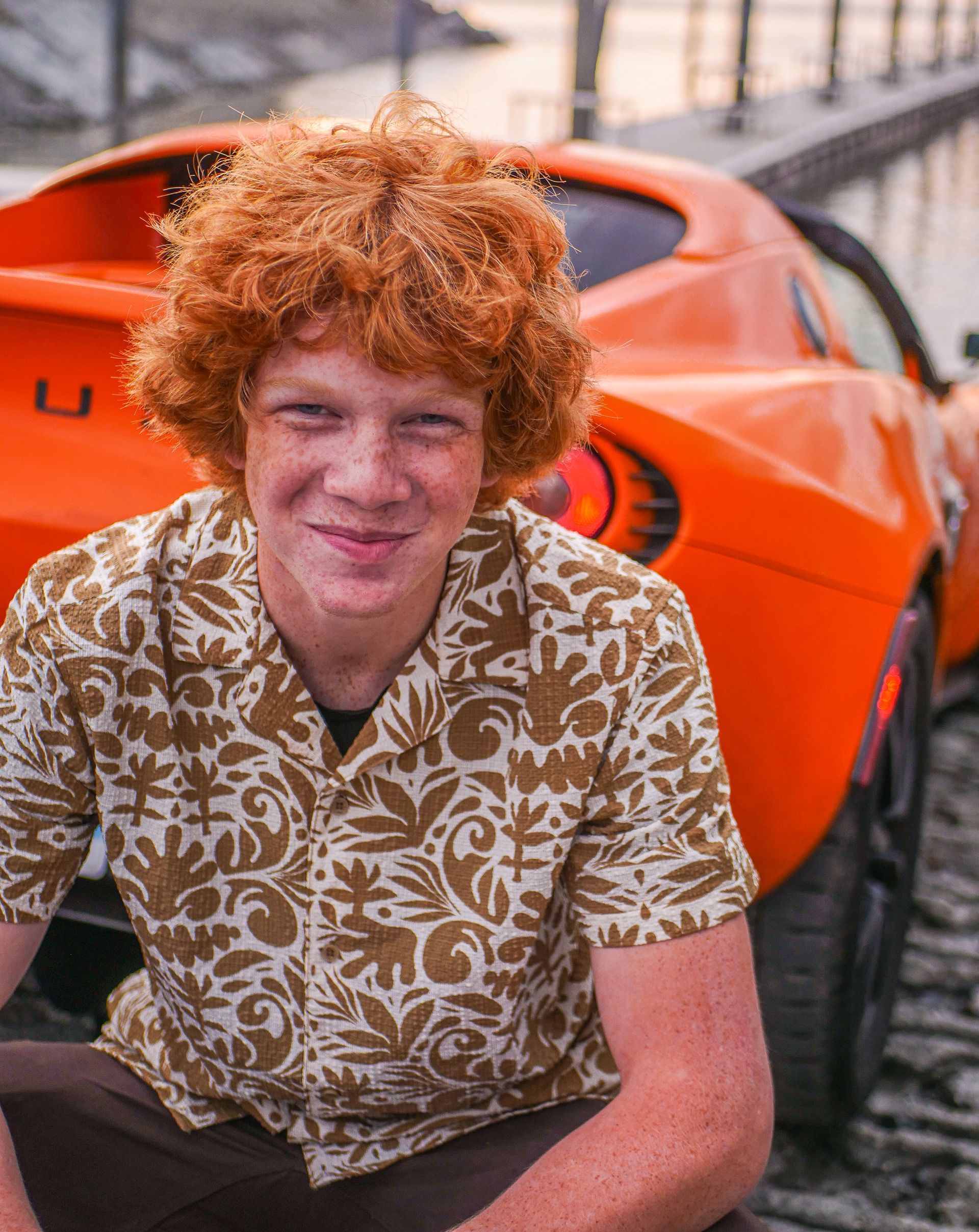 A young man with red hair is sitting in front of an orange sports car.