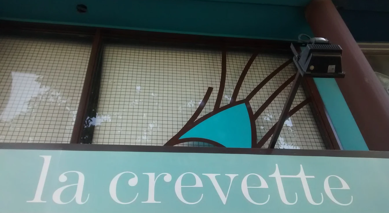 Letrero con la inscripción “la crevette” debajo de un toldo turquesa con diseño de ramas marrones.