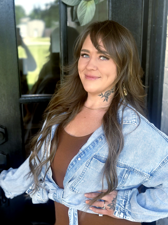 A person with long brown hair, wearing a brown top and light blue denim jacket, smiling at the camera near a doorway.