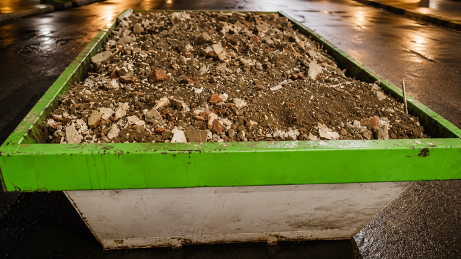 Un contenedor de basura verde lleno de tierra y escombros en una calle mojada por la noche.