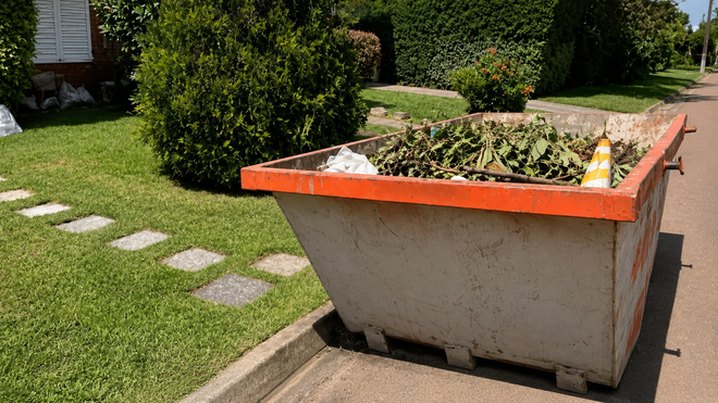 Un gran contenedor de obra gris con borde naranja se encuentra en un soleado jardín residencial cerca de losas y setos.