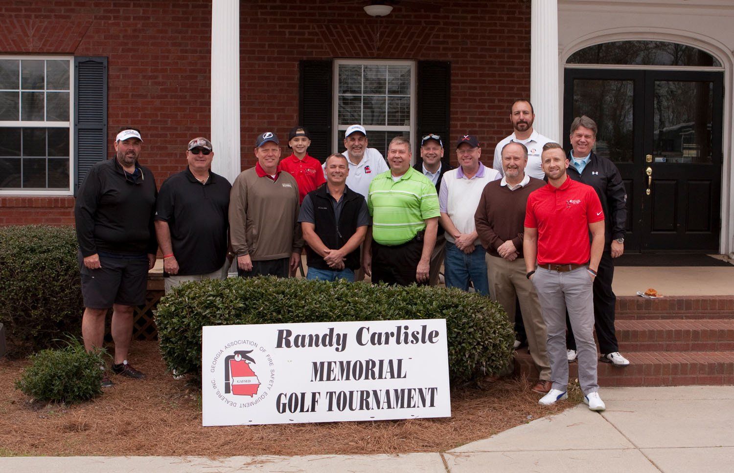 A group of men standing in front of a building with a sign that says randy carlisle memorial golf tournament