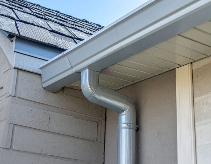Silver rain gutter and downspout on a light brown house with a dark gray roof.