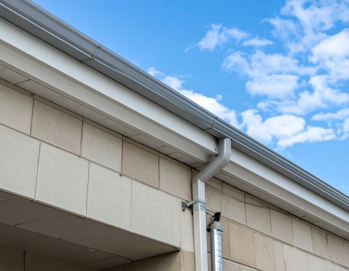Gray gutter and downspout on a light-colored brick building against a blue sky with clouds.