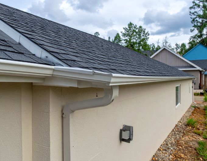 Beige building with dark gray roof, white gutters, and a downspout against a cloudy sky.