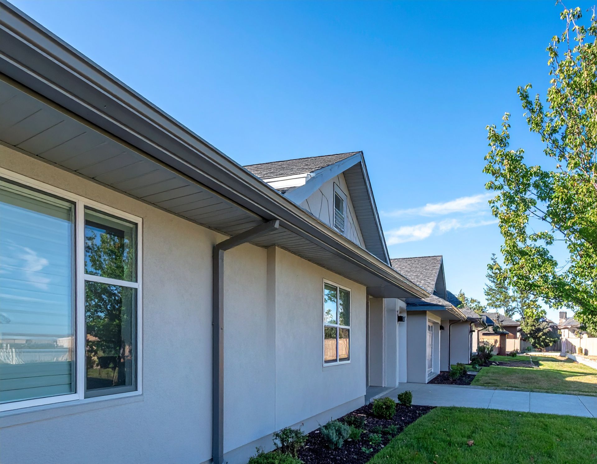 Suburban house exterior with beige siding, gray roof, and a sunlit green lawn under a blue sky