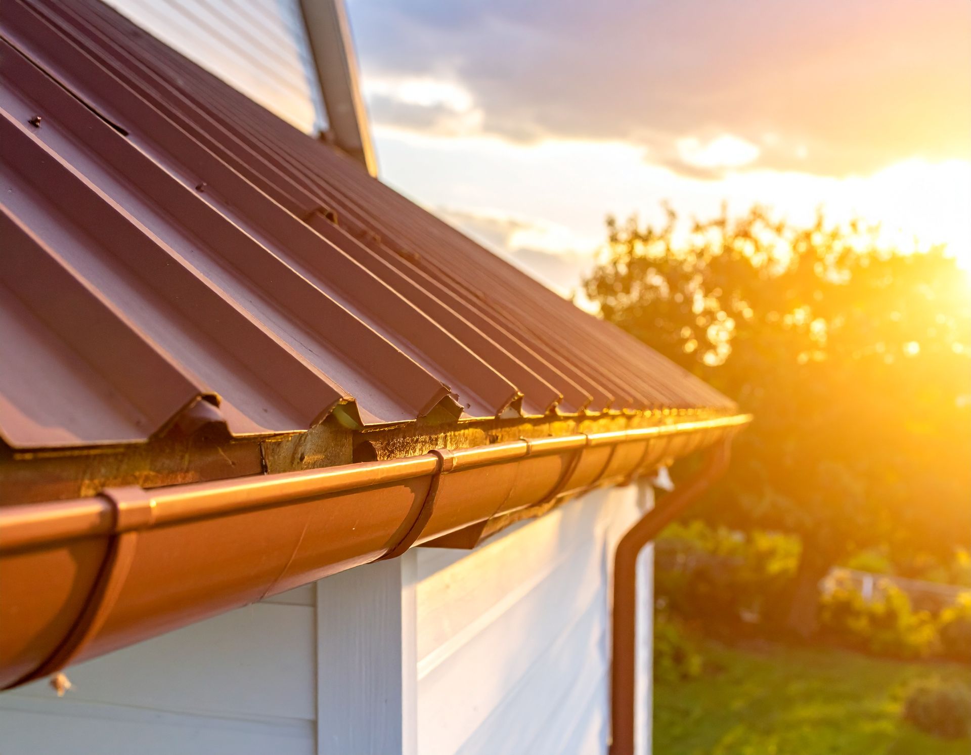 Sunlit house roof with brown metal panels and gutter at sunset