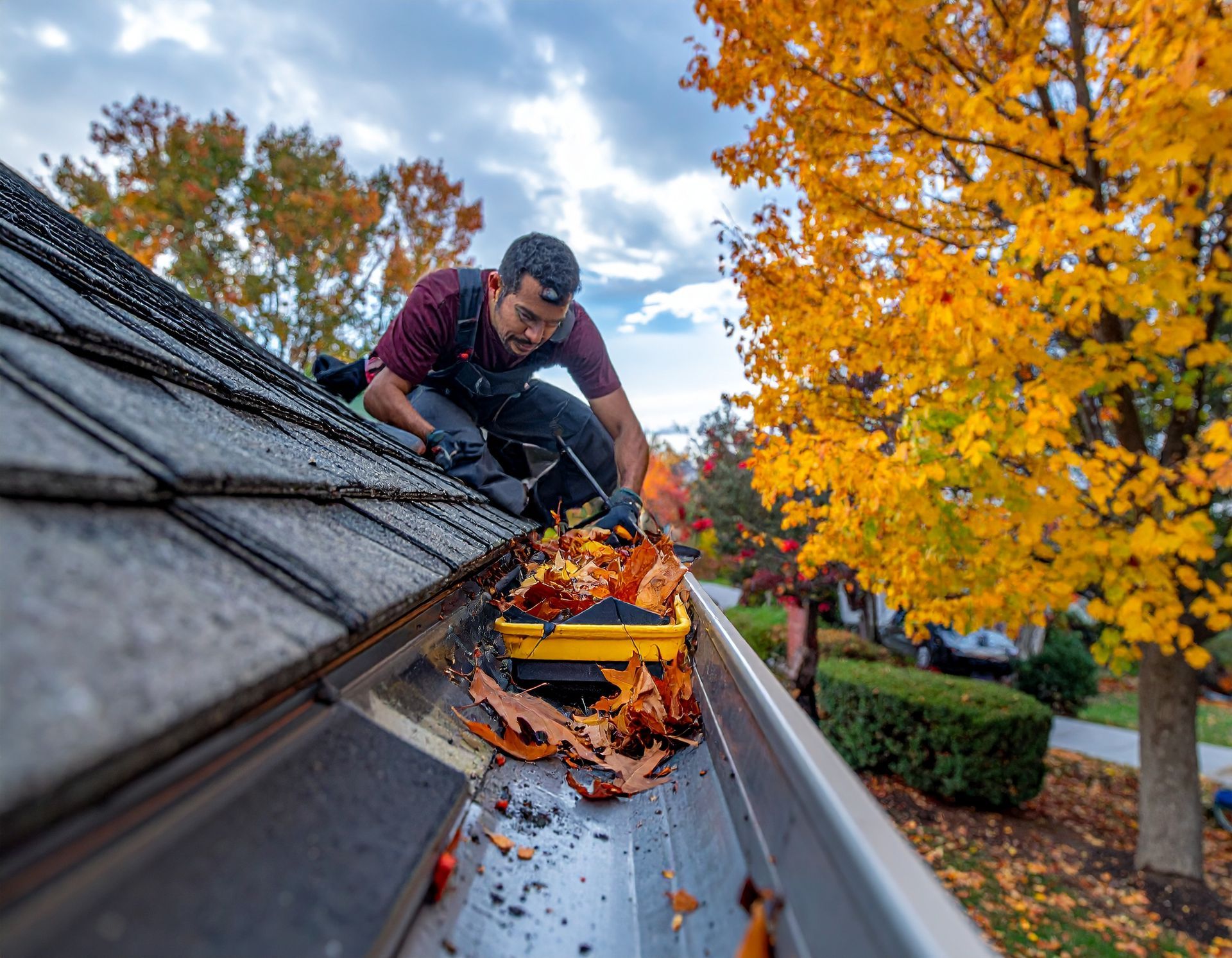 Worker cleaning a roof gutter filled with autumn leaves beside a bright orange tree